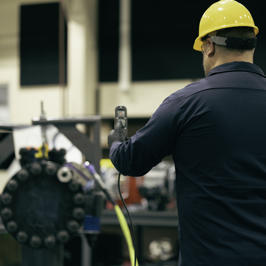 A technician operating a STEALTH Hydraulic Torque Wrench from a distance. He is wearing a hard hat, safety gloves and glasses, and using a remote pendant.