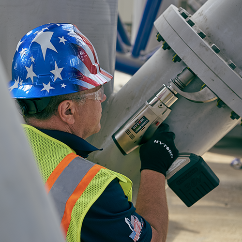 A technician wearing a USA-themed hard hat and safety glasses, holding a LITHIUM Series II Electric Torque Tool onto an application.