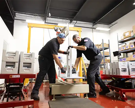 Two technicians are seen placing weights on a master calibration system intended to calibrate transducers in line with ISO/IEC 17025 standards and verify their accuracy.